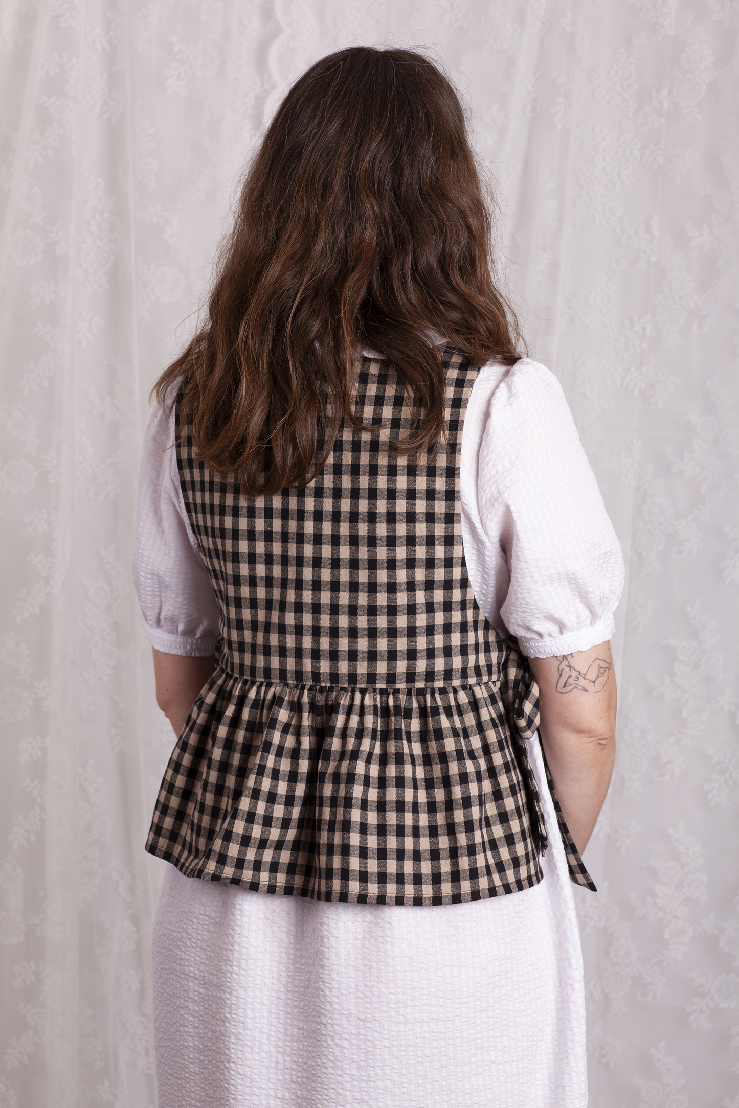 Back view of woman wearing a white dress and black and beige gingham Poppy Apron Top with a plain background. Buy the Poppy Apron Top PDF sewing pattern. 