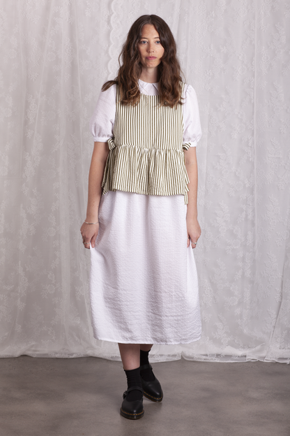 Woman wearing a white dress and green stripe Poppy Apron Top with a plain background. Buy the Poppy Apron Top PDF sewing pattern. 