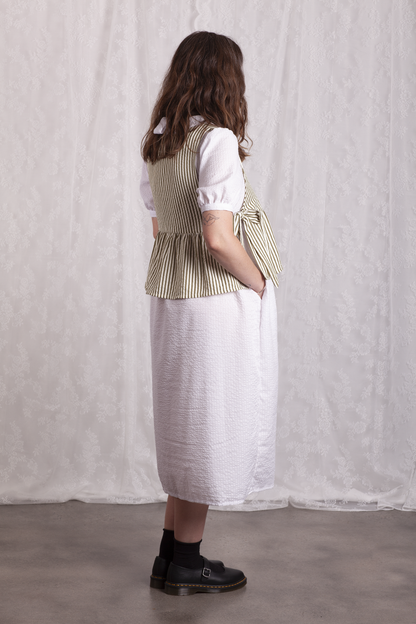 Side view of woman wearing a white dress and green stripe Poppy Apron Top with a plain background. Buy the Poppy Apron Top PDF sewing pattern. 