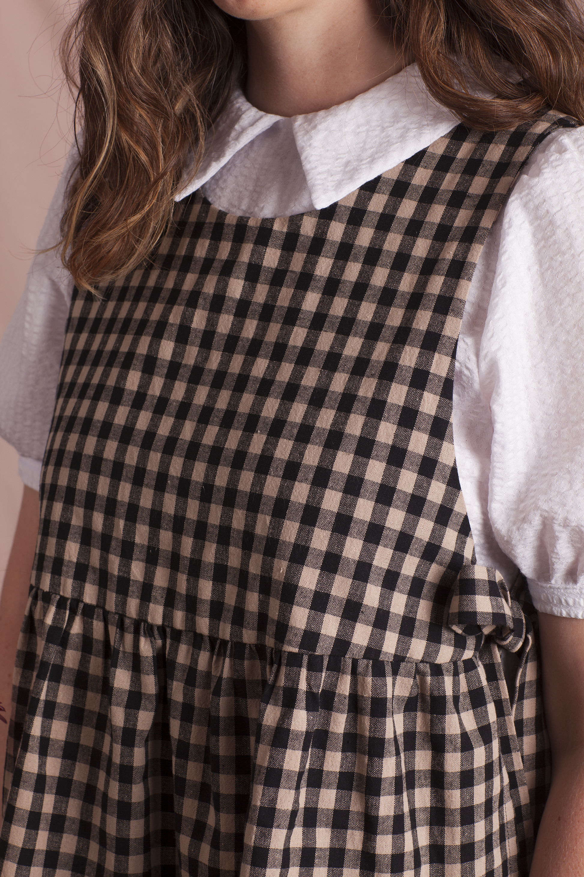 Close up of woman wearing a white dress and black and beige gingham Poppy Apron Top with a plain background. Buy the Poppy Apron Top PDF sewing pattern. 